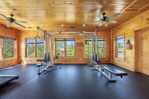 Ein Fitnessraum mit Deckenventilatoren, Trainingsgeräten und großen Fenstern mit Blick auf Bäume.