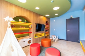 Ein Kinderspielzimmer mit Zelt, roten Hocker, Regalen voller Spielzeug, einem Fernseher und einer Tür mit Ausgangsschild.
