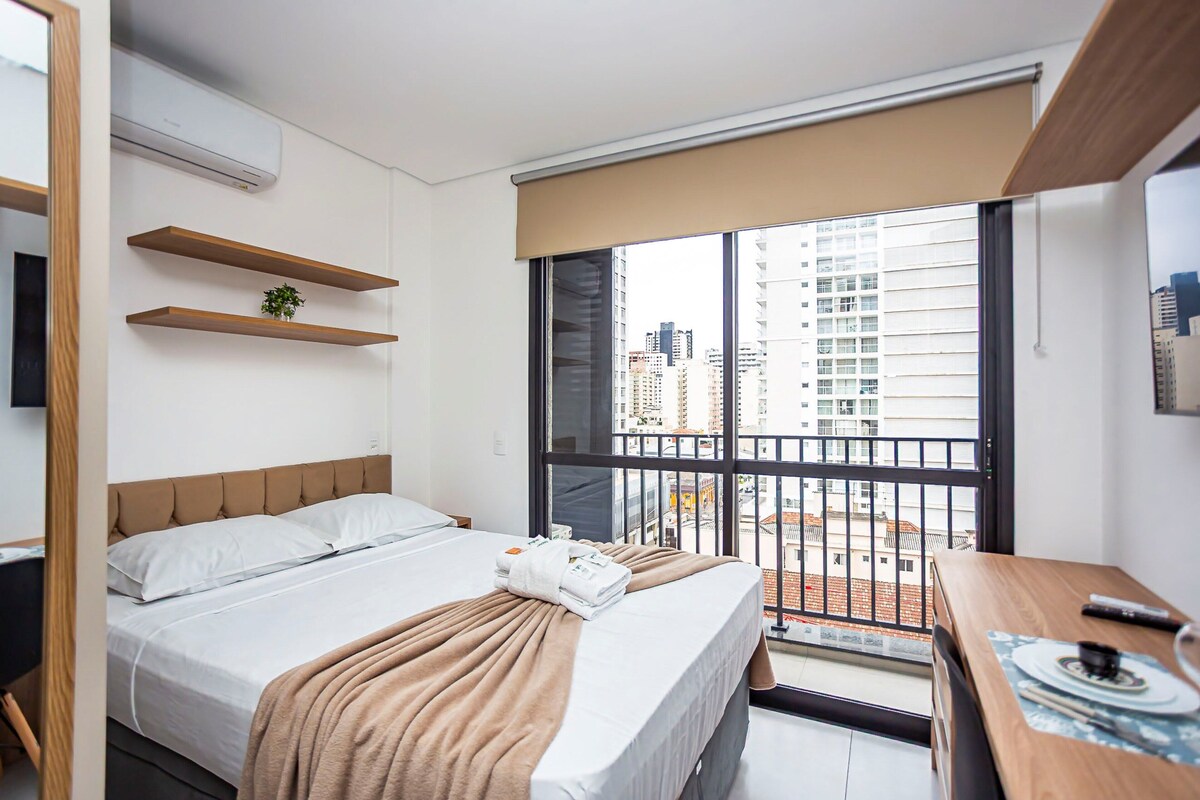 A hotel room with a bed, a desk, a chair, a window with a view of buildings, and a balcony.