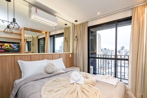 A hotel room with a bed, a large window, an air conditioning unit, and a city view.