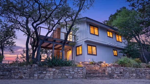 A two-story house with a stone wall, wooden deck, and a staircase leading to the entrance. - Lakefront Home w/Views, Huge Deck, Family-Friendly (Lago Vista)