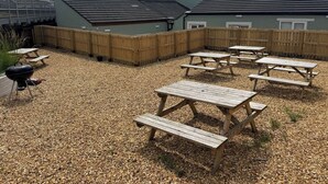 Een achtertuin met picknicktafels, een barbecue en een houten omheining.