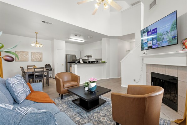 A modern living room with a sofa, chairs, a coffee table, a fireplace, a TV, and a kitchen in the background.