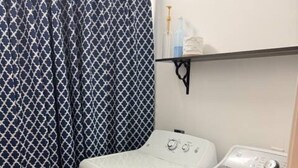 A laundry room with a washing machine and dryer, a patterned curtain, and a shelf with bottles. - Delightful 1-bedroom poolside house with AC in charming Hahira. (Hahira)