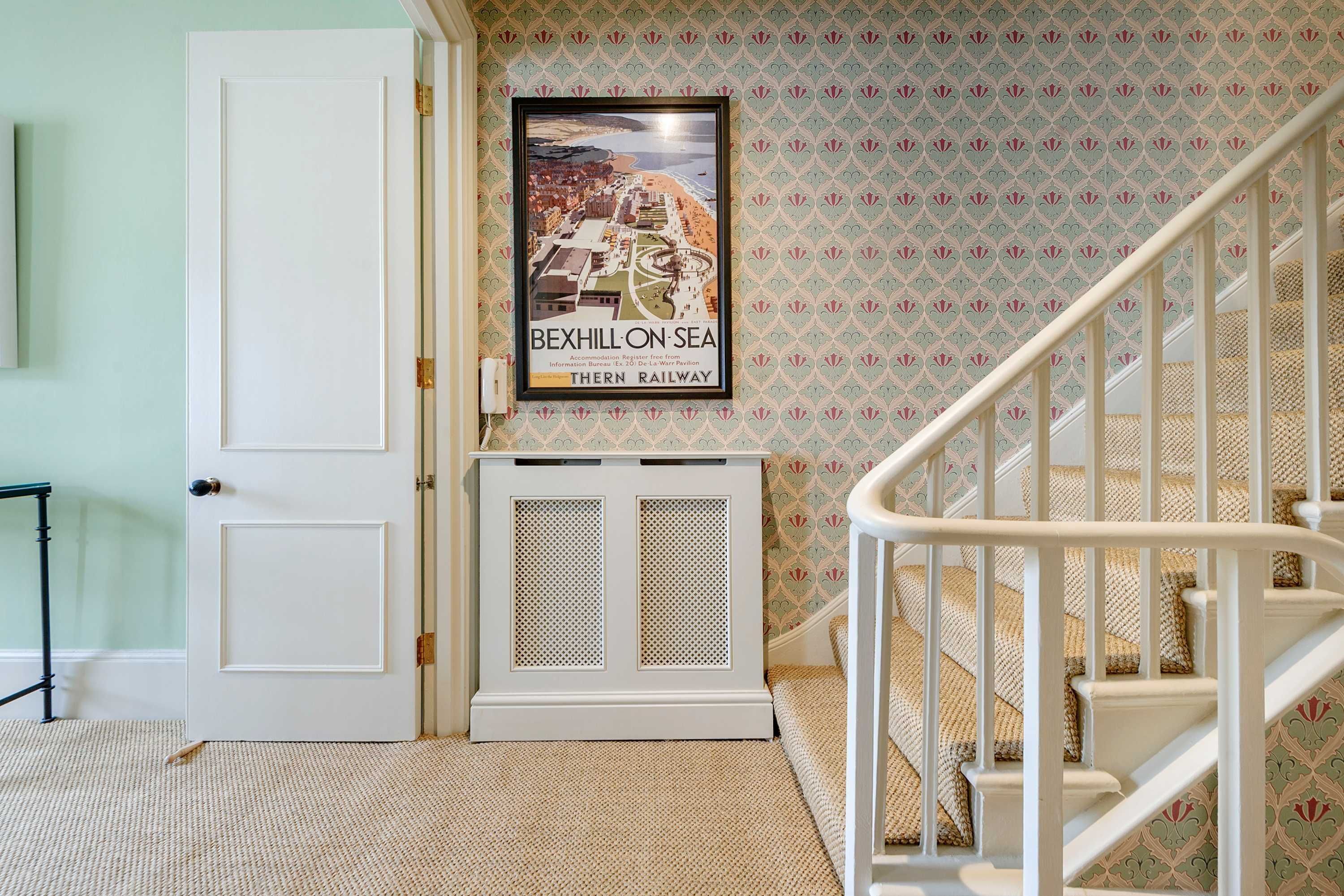 A staircase with a white railing, a framed poster of Bexhill-on-Sea, and a white door with a small table beside it.