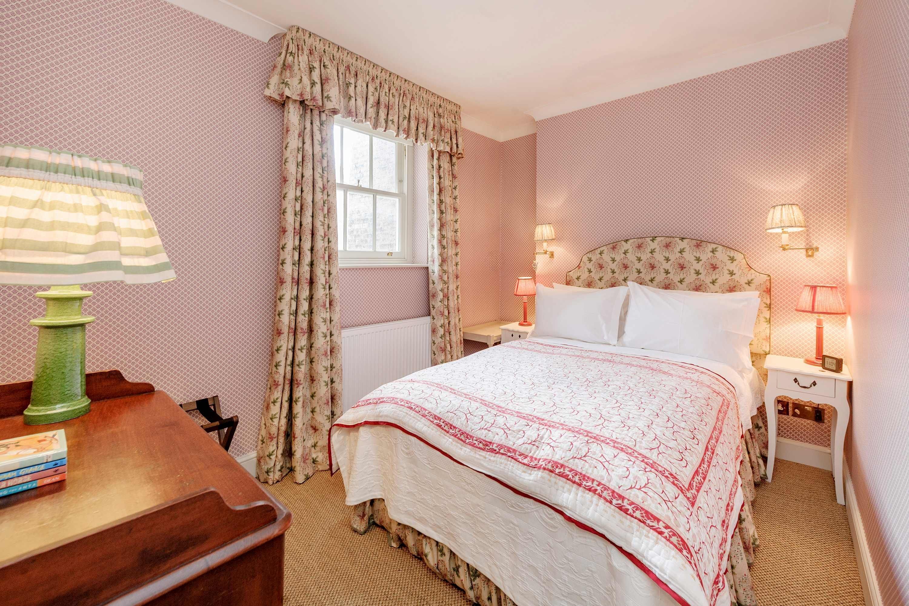 Een slaapkamer met een bed, twee nachtkastjes met lampen, een raam met gordijnen, een radiator en een houten bureau met boeken.