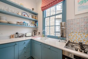 A kitchen with blue cabinets, a white countertop, a window with a red valance, and a patterned backsplash. - Gorgeous 3 Bedroom Flat - Pimlico (London)