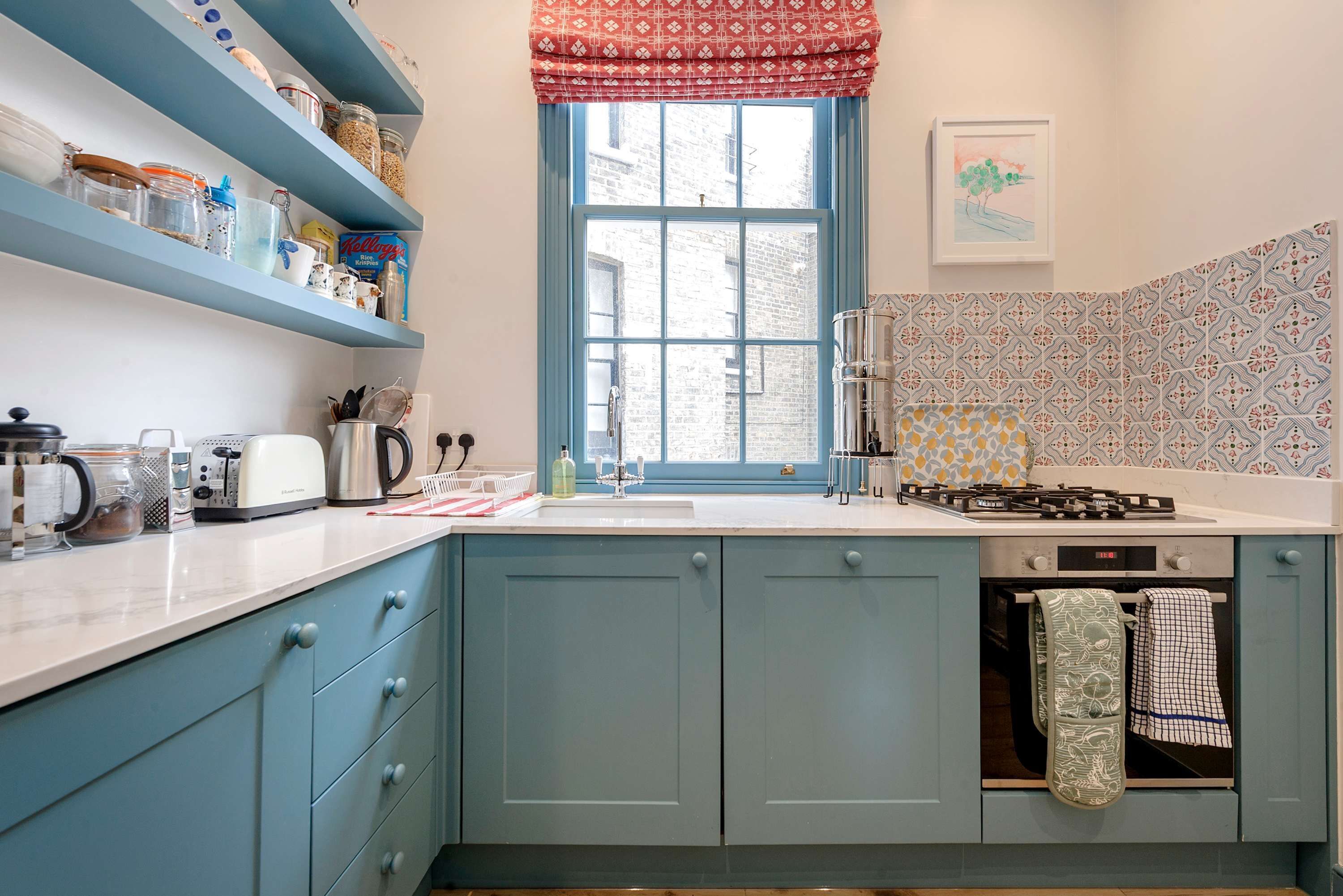 Een keuken met blauwe kastjes, een raam met een rode lambrizzen, een gasfornuis en een gootsteen.