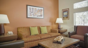 A hotel room with a sofa, armchair, coffee table, lamps, a telephone, and two framed pictures on the wall.