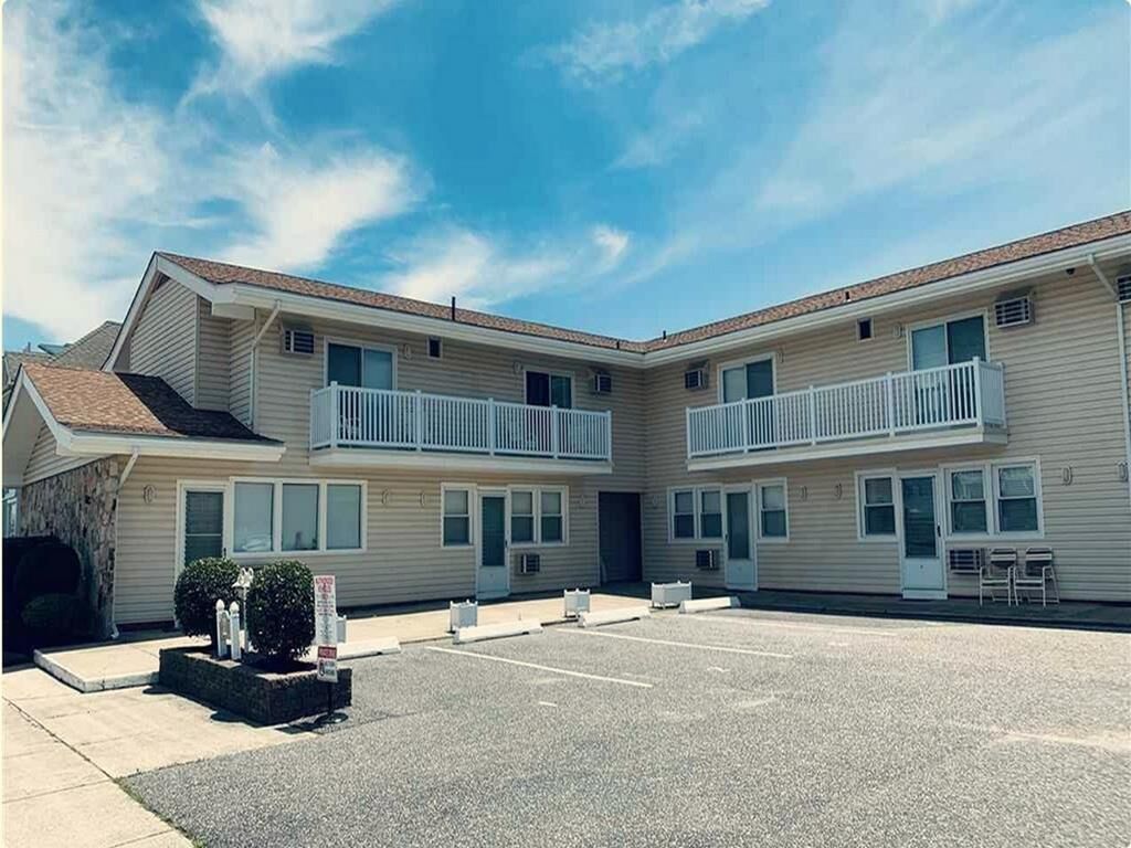 Ein zweistöckiges Hotel mit Parkplatz davor, einem kleinen Garten und blauem Himmel.