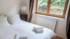 En snyggt bäddad säng med vita lakan och två hoprullade handdukar, en sänglampa, ett fönster med gardiner och ett element.