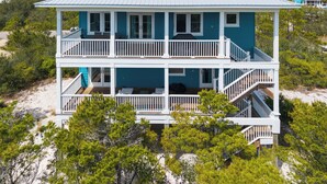 Una casa de playa de dos pisos con balcones, techo de metal y rodeada de árboles y arena.