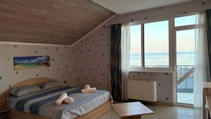 A bedroom with a bed, a wooden headboard, a view of the sea, a window with curtains, and a small table. - Vista Mare Sveti Vlas (Sveti Vlas)