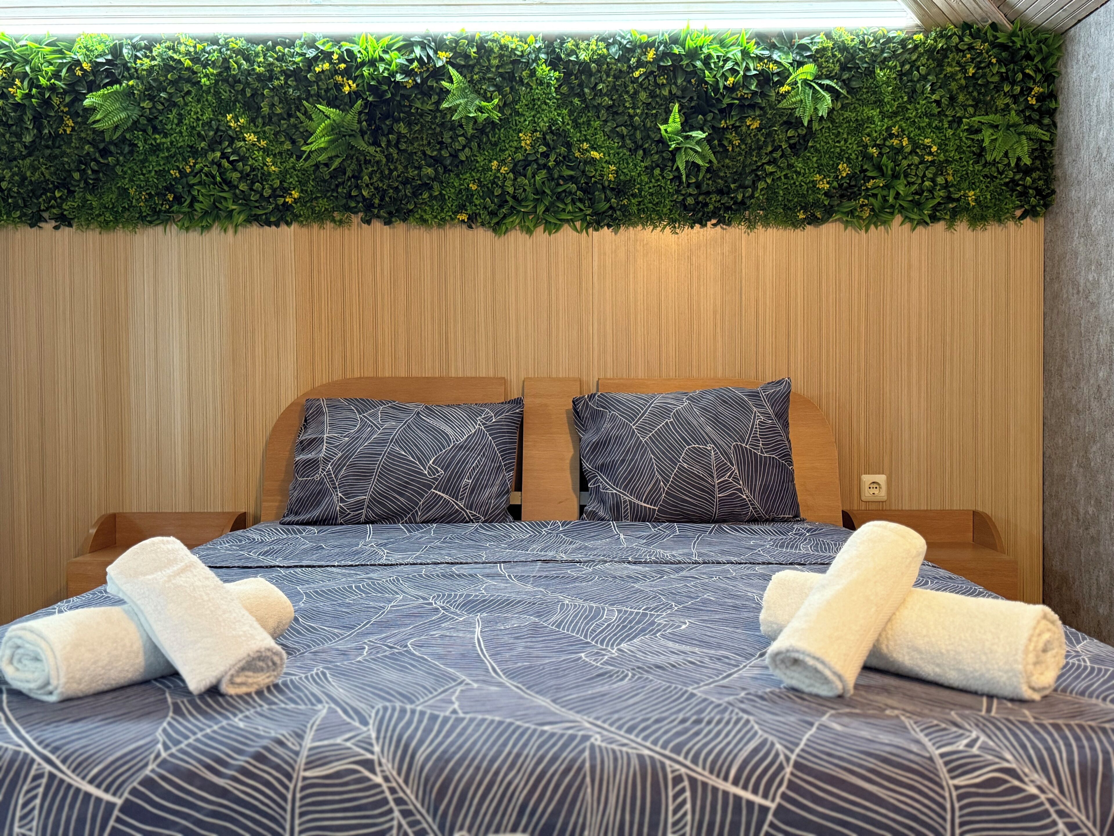 Een bed met een beddengoed met bladmotief, twee kussens en opgerolde handdoeken op een houten hoofdbord, met een groene plantenwand erboven.
