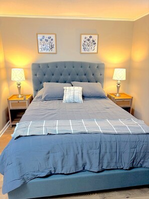 Ein Bett mit gebuschter Kopfteil, zwei Nachttische mit Lampen und zwei gerahmte Blumenbilder an der Wand.