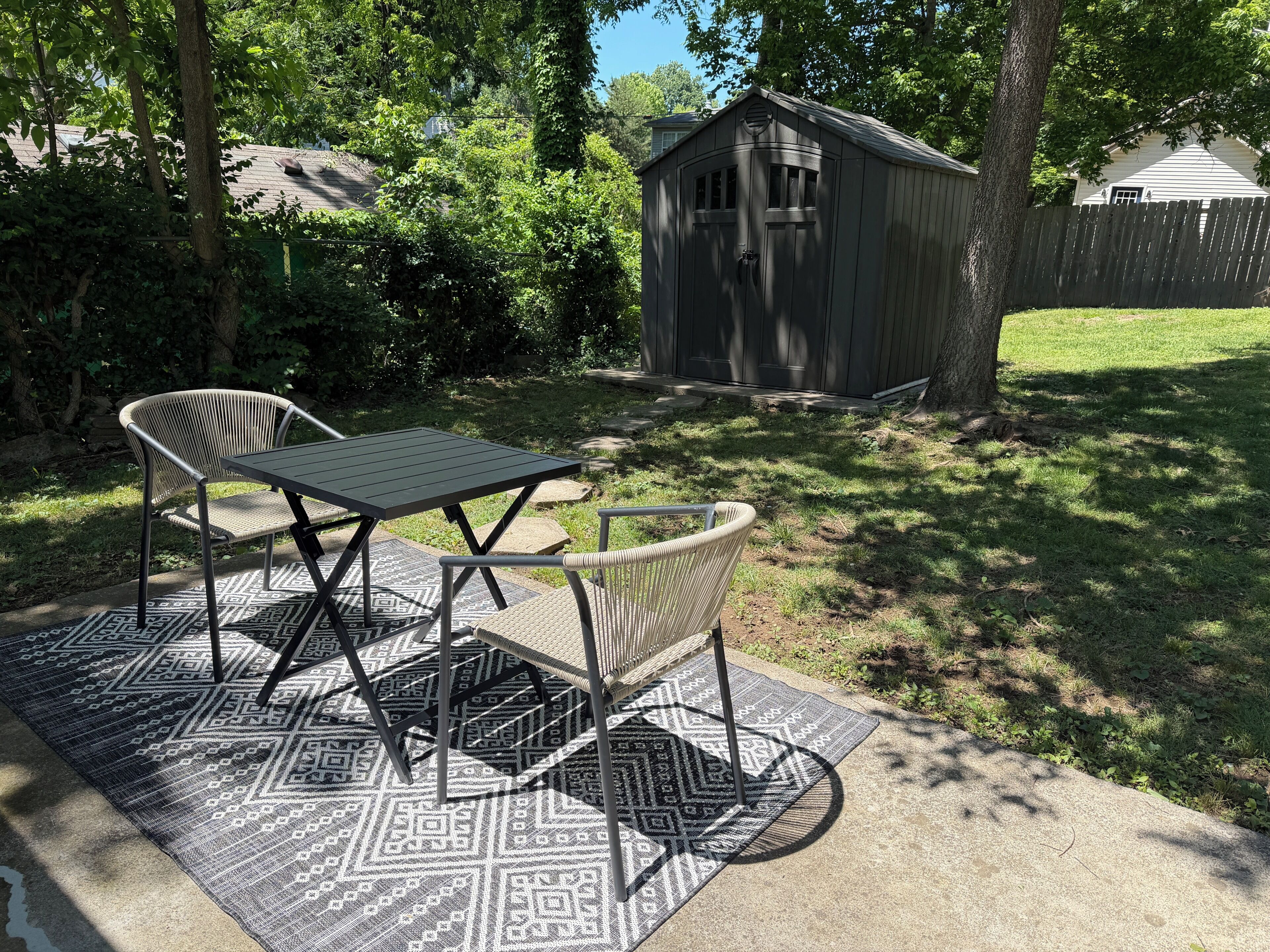 A patio with a table and chairs, a patterned rug, a shed, and a wooden fence.
