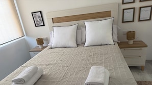 Un lit bien fait, recouvert d’une couvre-lit à motifs, avec deux oreillers et deux serviettes enroulées. Des tableaux encadrés ornent le mur, et une table de chevet avec une lampe se trouve à côté du lit.