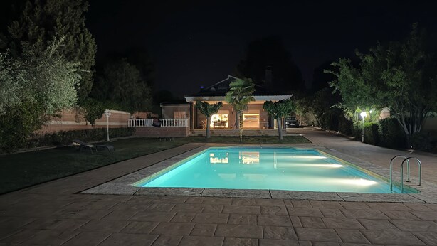 Une piscine rectangulaire entourée d’une terrasse dallée, bordée d’arbres, et en arrière-plan, un bâtiment doté d’un patio couvert.
