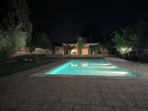 Une piscine rectangulaire entourée d’une terrasse dallée, bordée d’arbres, et en arrière-plan, un bâtiment doté d’un patio couvert.
