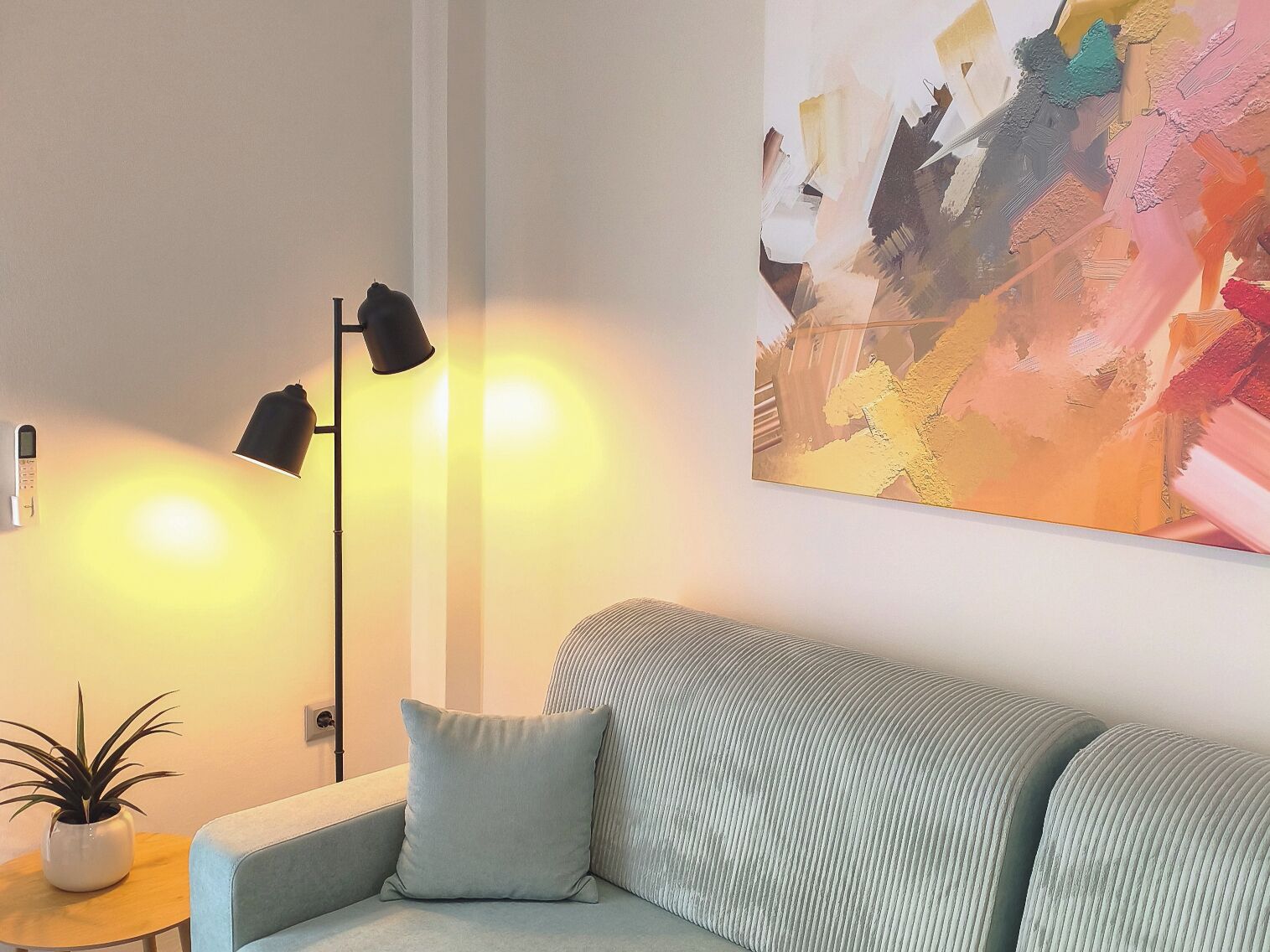 A modern living room with a sofa, a lamp, a small table with a plant, and a colorful abstract painting on the wall.
