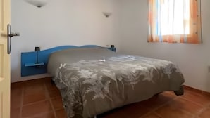 A bedroom with a bed, a blue headboard, a wall-mounted shelf, and a window with curtains.