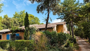 A rustic house with a tiled roof, surrounded by dense greenery and a gravel path.