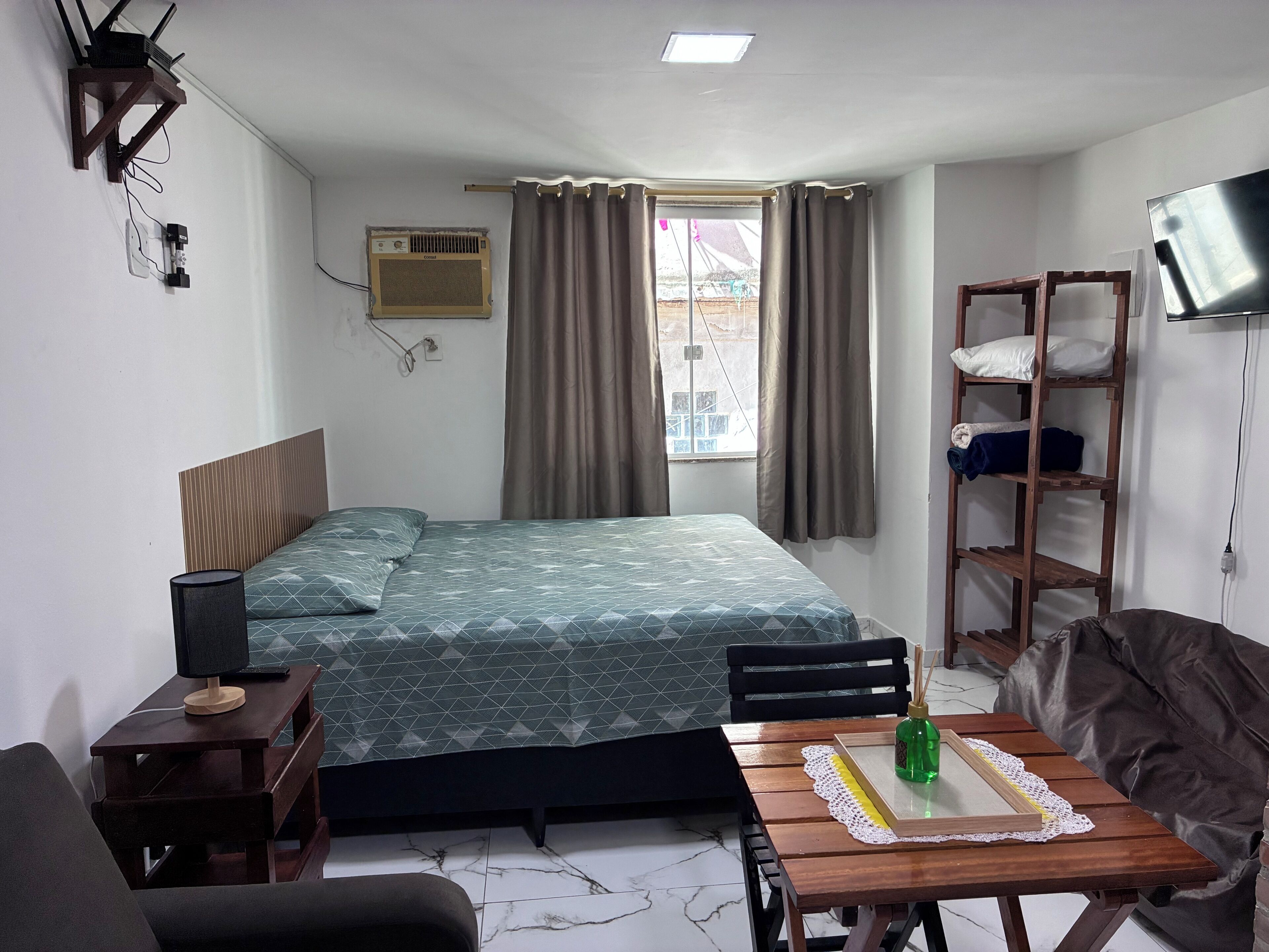 Quarto com cama, mesa de cabeceira, luminária, televisão e janela com cortinas.