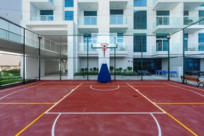 Eine Basketballhalle mit einem blauen Korb, umgeben von einem Zaun, und ein Gebäude mit zahlreichen Fenstern im Hintergrund.