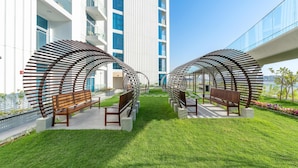 A modern outdoor seating area with benches under curved metal structures, surrounded by a well-maintained lawn and a building with balconies. - Free Housekeeping Visits During Your Stay - Stayshort - Modern New Studio in Arjan That Sleeps 4 (Dubai)