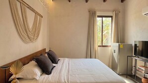 A bedroom with a bed, wooden ceiling beams, a ceiling fan, a window with curtains, a television, and a refrigerator. - Casa Florentina (Tulum)