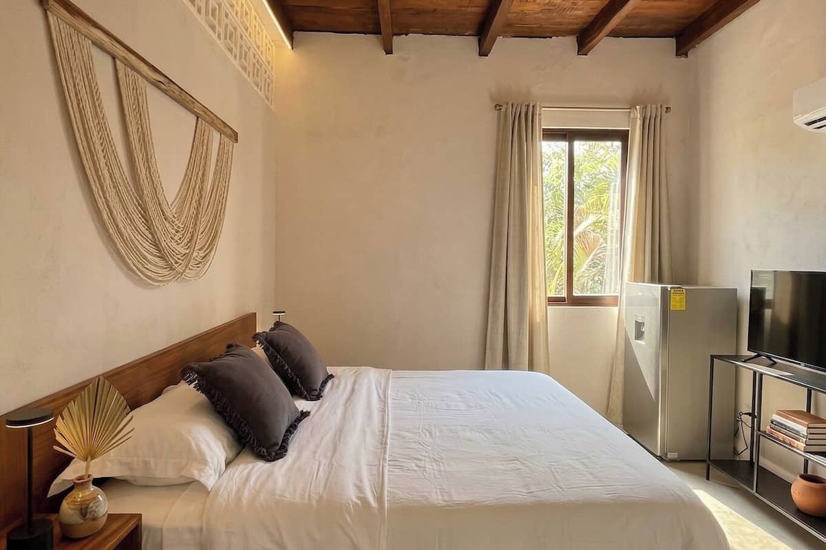 A bedroom with a bed, wooden ceiling beams, a ceiling fan, a window with curtains, a television, and a refrigerator.
