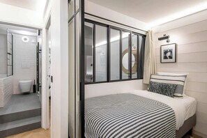 Un dormitorio moderno con cama, una ventana con persianas, un cuadro en la pared y un baño a la vista al fondo.
