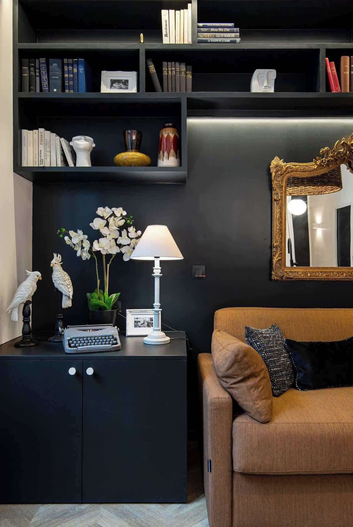 Un salon moderne avec une bibliothèque noire, une lampe blanche, un tableau encadré et un miroir doté d’un cadre orné.