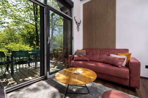 A modern living room with a sofa, coffee table, and a view of a forest through large glass doors.