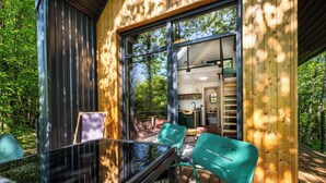 Eine Holzterrasse mit Glastisch und Stühlen, ein durch die Fenster einsehbarer Küchenbereich und Blick auf den umliegenden Wald.