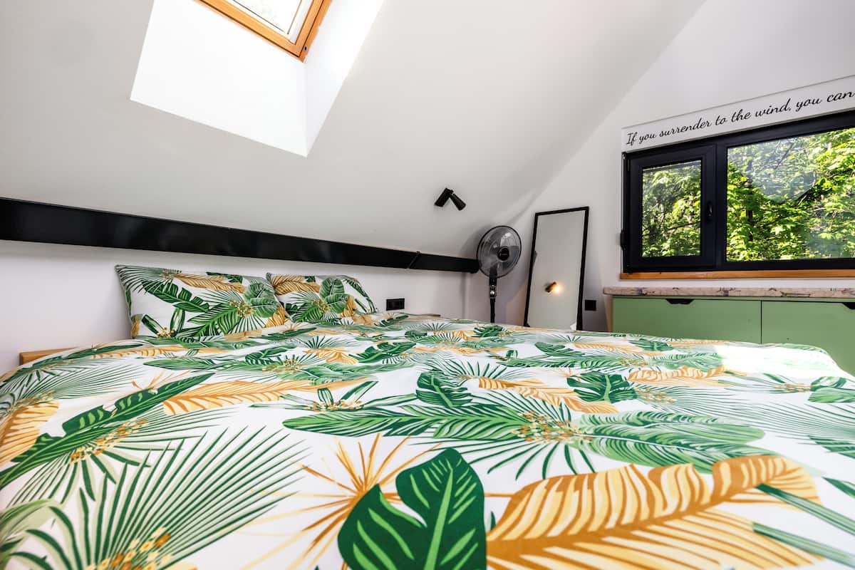 Une chambre à coucher avec un lit, une lucarne, une fenêtre donnant sur un beau paysage et un ventilateur fixé au mur.