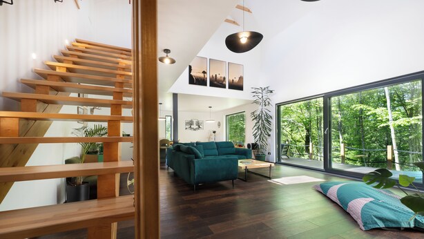 Ein modernes Interieur mit einer Holztreppe, einem türkisblauen Sofa und großen Fenstern mit Blick ins Grüne.