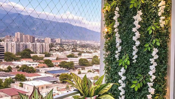 Un balcón con vistas a un paisaje urbano, una pared verde con flores blancas y plantas en macetas.