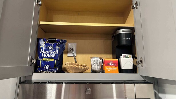 A kitchen with a coffee maker, a bag of Maxwell House coffee, a glass of water, and a box of Folgers breakfast blend.