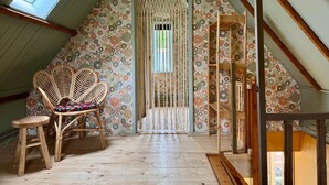 Ein Zimmer mit geneigter Decke, einer Tür mit dekorativem Vorhang, einem Korbstuhl, einem Holzschemel und einem Bücherregal aus Holz.