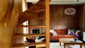 Eine Holztreppe führt zu einem erhöhten Bereich mit Backsteinwand, einer Couch, einem Couchtisch und einer Hängelampe.