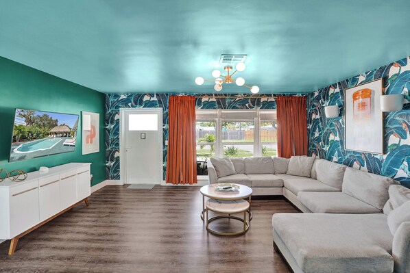 A living room with teal walls, a white sofa, a round coffee table, and a large window with curtains. - Tropical Pool Oasis · Heated Pool · 2BR · 2BA · Steps to Beach (Pompano Beach)