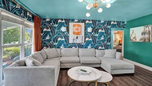 Una sala de estar con un sofá modular, una mesa de centro, un candelabro en el techo y un papel tapiz con motivos tropicales.