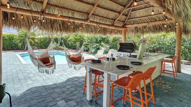 Un área de comedor al aire libre con techo de paja, piscina y parrilla.