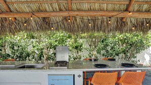 Una cocina con estufa integrada, un lavamanos y un refrigerador. Hay sillas anaranjadas y una mesa lista para comer.