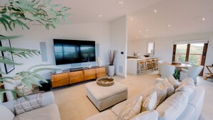 A spacious living area with a large flat-screen TV, a wooden console, a dining area, and a view of greenery.