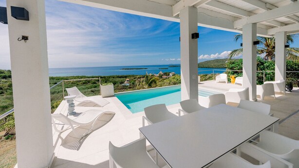 A covered outdoor area with a pool, white furniture, and a view of the ocean.