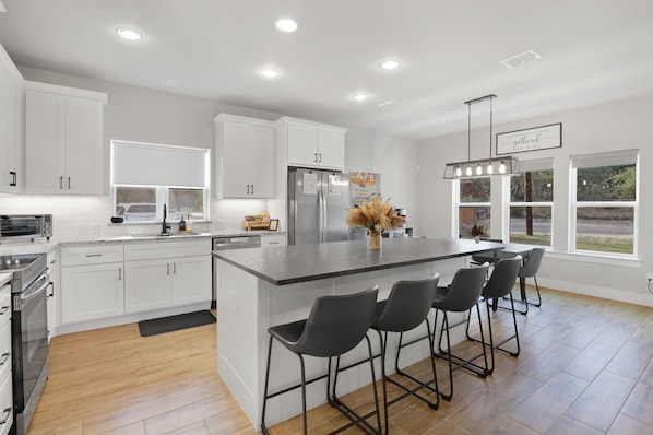 A modern kitchen with white cabinets, a central island with bar stools, a stainless steel refrigerator, and a hanging light fixture.