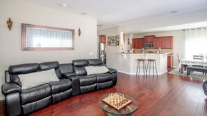 A living room with a black leather sofa, a glass coffee table with chess pieces, and a kitchen in the background. - 4BR/3BA, 3700 SqFt, Luxury Home in Durham near RDU, Duke, and UNC (Durham)