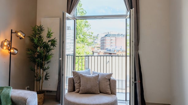 Ett rum med en soffa, en krukväxt, en lampa och en utsikt över byggnader genom ett fönster.